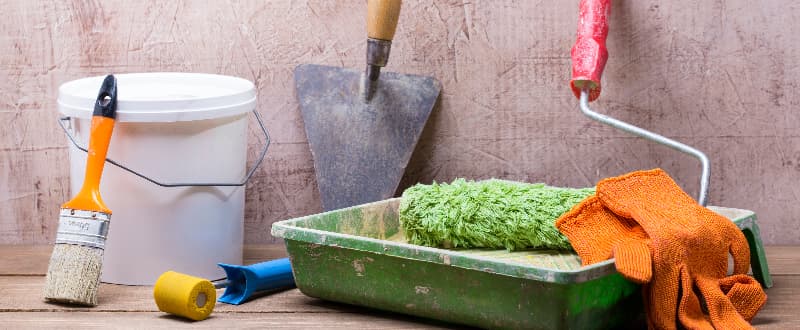 A collection of painting and decorating tools arranged on a wooden surface, including a paintbrush, white paint bucket, metal trowel, paint roller with green cover, green paint tray, orange cloth, and blue scraper, set against a textured wall background.